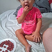 Nakaylia is registered to the contest to win money with this photo: toddler, child, grapes, food, bed, pink_shirt, person, sitting, indoor, funny_face, snack, plate, barefoot, furniture, home, curly_hair, expression, casual, light, clothing