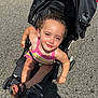 child, toddler, stroller, outdoor, sunlight, smiling, happy, colorful_clothing, footwear, sandals, pavement, black_stroller, person, summer, daylight, curly_hair, playful, seat, baby, casual