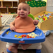 Kaiden joined the competition — help win amazing prizes! baby, high_chair, silicone_bib, food, corn, mashed_potatoes, tomatoes, floor, toys, book_shelf, books, door, carpet, child, indoor, smiling, furniture, feeding, playroom, person