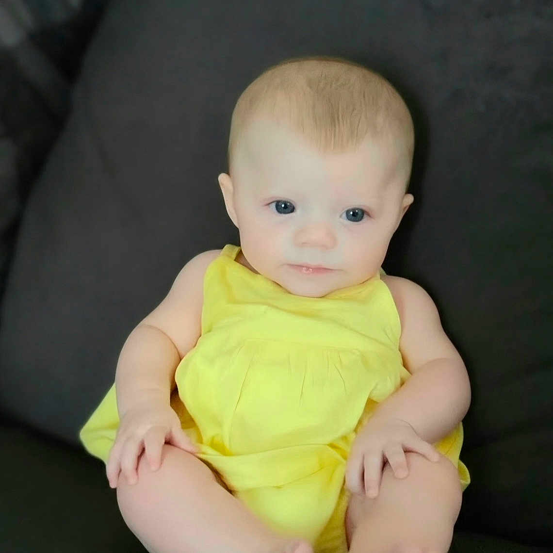 Kinsley joined the competition — help win amazing prizes! baby, infant, child, portrait, sitting, yellow_outfit, dress, dark_couch, indoor, blue_eyes, blond_hair, wide_eyed, barefoot, smiling, looking_at_camera, centered, closeup, soft_lighting, photograph, watermark