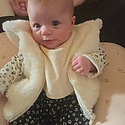 Kinsley is registered to the contest to win money with this photo: baby, infant, child, portrait, closeup, face, wide_eyed, sitting, fluffy_jacket, cream_clothing, floral_outfit, blanket, bed, cushion, stuffed_toy, indoors, cozy, looking_at_camera, hand, soft_lighting