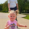 toddler, child, smiling, outdoor, sunny, pigtails, pink_shirt, text_on_clothing, adult, blurred_background, pathway, greenery, daylight, person, happy, casual_clothing, portrait, nature, playful, cute