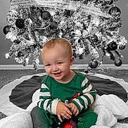 Kristopher is registered to the contest to win money with this photo: baby, child, christmas_tree, ornament, holiday, smiling, pajamas, blanket, basket, carpet, decorations, festive, indoor, celebration, joyful, cute, holiday_lights, red_ornament, green_clothing, smile