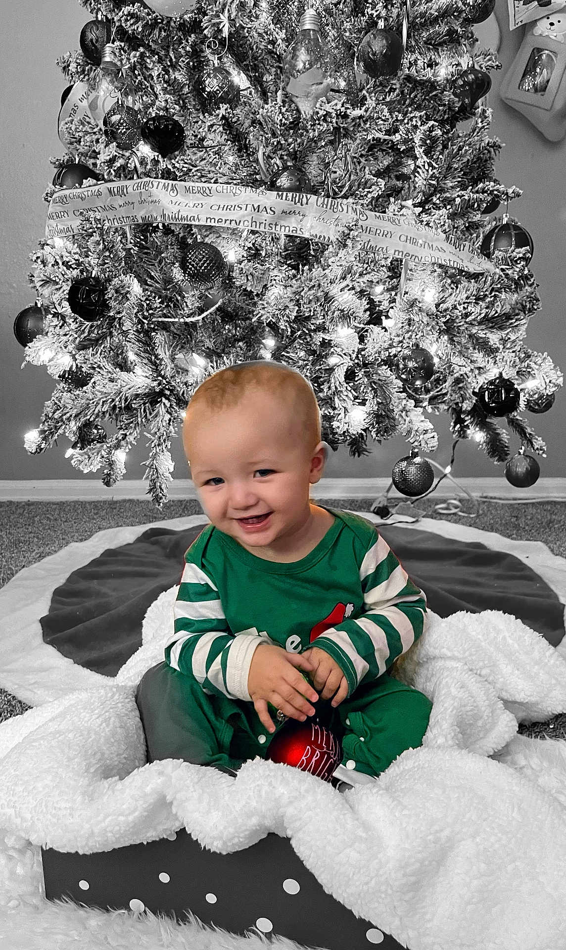 Kristopher is registered to the contest to win money with this photo: baby, child, christmas_tree, ornament, holiday, smiling, pajamas, blanket, basket, carpet, decorations, festive, indoor, celebration, joyful, cute, holiday_lights, red_ornament, green_clothing, smile