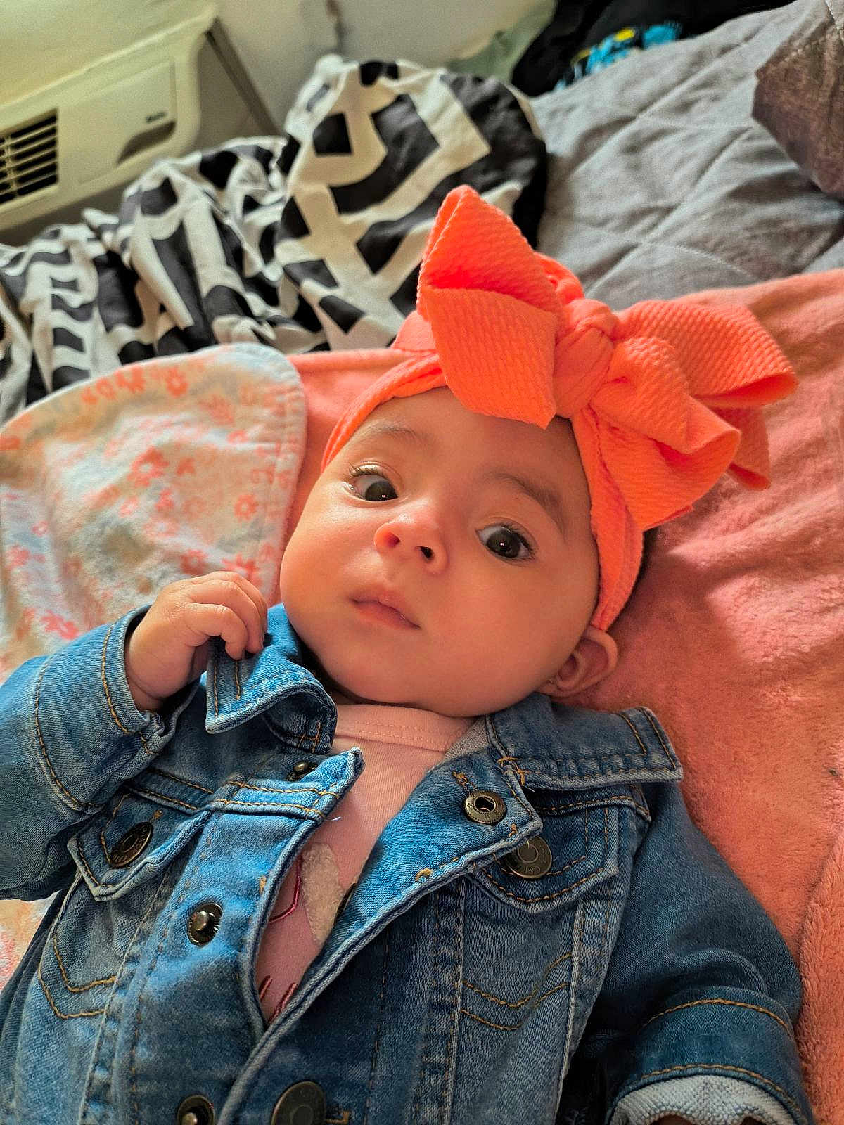 Aislynn is registered to the contest to win money with this photo: baby, denim_jacket, orange_bow, headband, blanket, indoor, infant, cute, face, portrait, child, clothing, soft_texture, lying_down, curious, young, person, cozy, patterned_blanket, casual