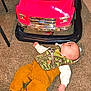 Kolton is registered to the contest to win money with this photo: baby, infant, carpet, toy_car, pink_car, ride_on_toy, chrome_grille, bumper, camouflage_bib, bib, brown_pants, leather_boots, lying_down, looking_up, indoor, floor, vehicle, clothing, hand, face