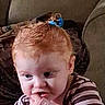 toddler, child, red_hair, striped_shirt, blue_hair_clip, couch, blanket, indoor, person, young_child, curious, finger_in_mouth, box, pink, brown_furniture, seated, casual_clothing, portrait, home, expression