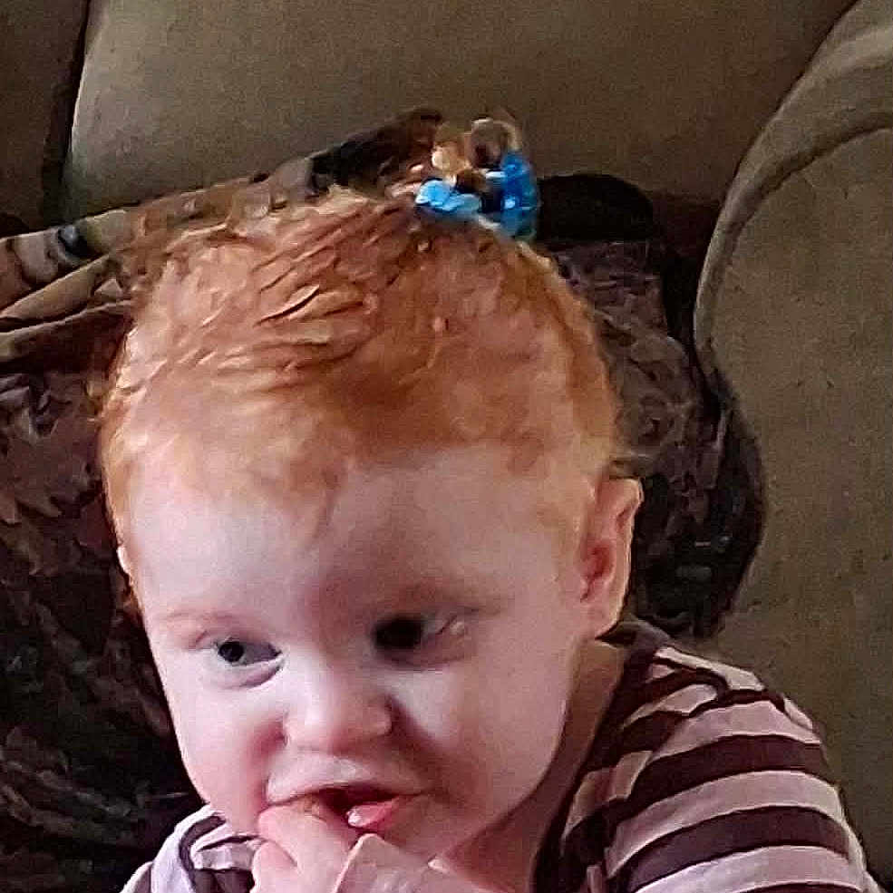 Blair joined the competition — help win amazing prizes! toddler, child, red_hair, striped_shirt, blue_hair_clip, couch, blanket, indoor, person, young_child, curious, finger_in_mouth, box, pink, brown_furniture, seated, casual_clothing, portrait, home, expression