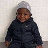 child, toddler, smiling, sitting, black_tracksuit, nike_logo, beanie, gray_hat, happy, indoor, floor, wall, portrait, cute, young_child, fashion, casual_wear, person, clothing, headwear