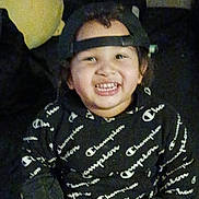 Jay'Quan is registered to the contest to win money with this photo: child, smiling, hoodie, cap, backwards_cap, sitting, indoor, couch, pillow, happy, person, face, clothing, casual_wear, young, portrait, cute, black_hoodie, text_pattern, cheerful