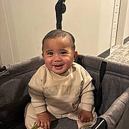 Duke is registered to the contest to win money with this photo: baby, smiling, child, stroller, indoor, clothing, foot, sitting, face, happy, person, apparel, infant, cute, young, cozy, warm, expression, portrait, furniture