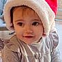 toddler, child, santa_hat, smiling, indoor, window, soft_lighting, cute, holiday, festive, portrait, baby, face, clothing, gray_dress, hands, happy, young_child, cozy, closeup