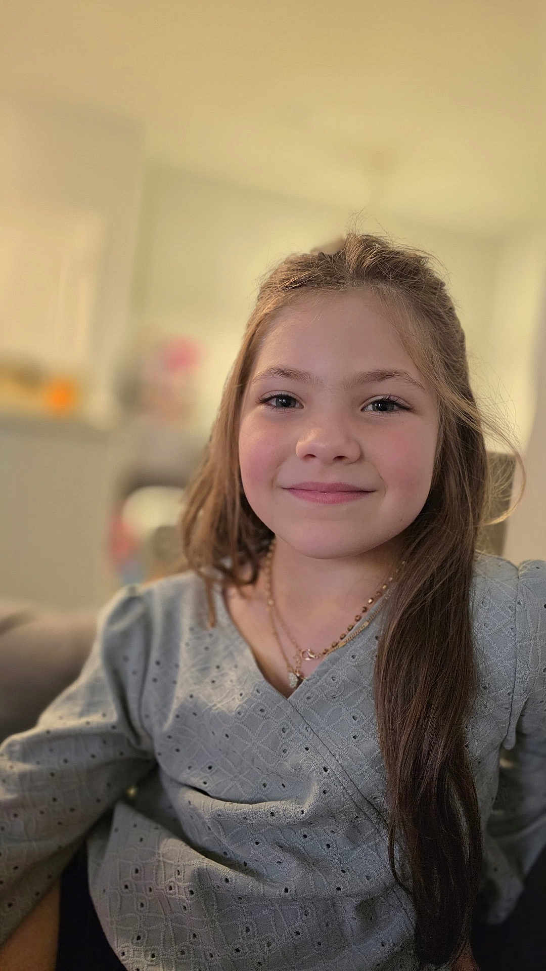Sophie is registered to the contest to win money with this photo: child, girl, smile, long_hair, necklace, gray_top, indoor, portrait, couch, happy, casual_clothing, person, home, young, face, seated, relaxed, fashion, lifestyle, blurry_background