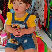 Bella is registered to the contest to win money with this photo: toddler, child, yellow_shirt, denim_overalls, indoor, clothing, washing_machine, sippy_cup, blanket, curly_hair, holding_object, colorful_clothes, laundry_room, cute, portrait, seated, baby, casual, home, expression