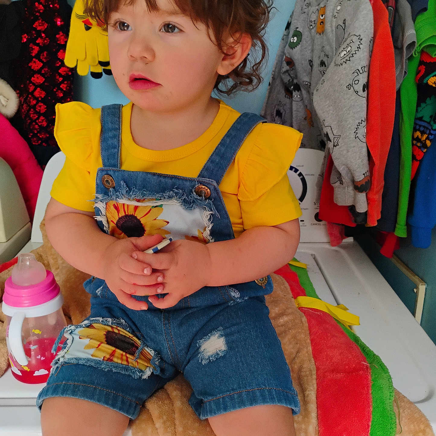 Bella is registered to the contest to win money with this photo: baby, blanket, casual, child, clothing, colorful_clothes, curly_hair, cute, denim_overalls, expression, holding_object, home, indoor, laundry_room, portrait, seated, sippy_cup, toddler, washing_machine, yellow_shirt