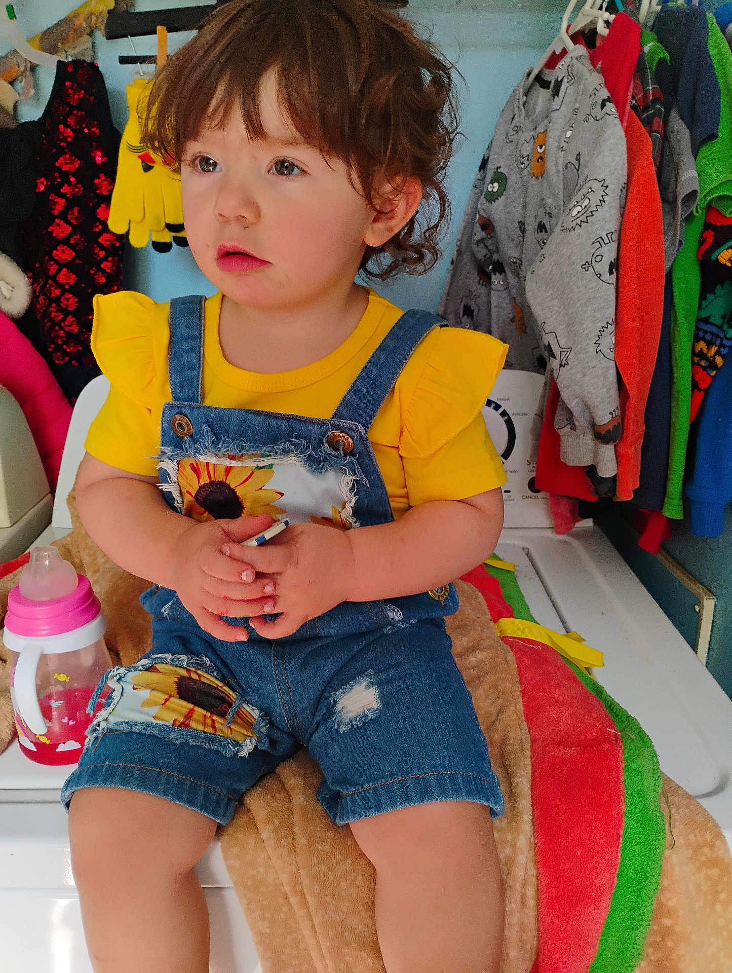 Bella is registered to the contest to win money with this photo: toddler, child, yellow_shirt, denim_overalls, indoor, clothing, washing_machine, sippy_cup, blanket, curly_hair, holding_object, colorful_clothes, laundry_room, cute, portrait, seated, baby, casual, home, expression