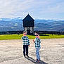 child, children, boys, sunglasses, plaid_shirts, matching_outfits, peace_sign, tower, mountains, scenic, grass, picnic_benches, fence, sky, clouds, shadow, pavement, daylight, outdoors, pose