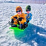 child, two_children, young_child, sled, snow, winter_clothing, beanie, sunglasses, gloves, boots, blue_sky, snowy_field, red_fence, outdoor, shadow, play, fun, winter_activity, sitting, bright_day