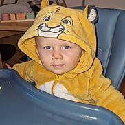 Nolan a rejoint le concours — aidez-le/la à gagner de superbes lots ! baby, child, high_chair, yellow_hoodie, lion_costume, face, blue_eyes, indoor, table, pumpkin, toy, furniture, person, hood, cute, expression, zipper, seat, room, soft
