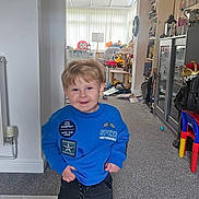 Radley joined the competition — help win amazing prizes! child, boy, blue_sweatshirt, standing, indoor, carpet, toys, furniture, smiling, living_room, cabinet, books, colorful_chair, radiator, patches, playroom, happy, hair, person, casual_clothing