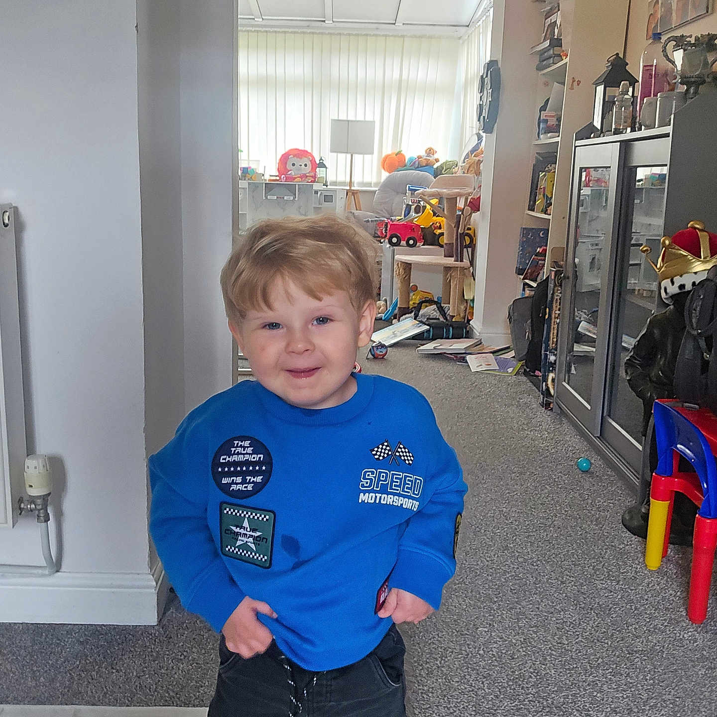 Radley joined the competition — help win amazing prizes! child, boy, blue_sweatshirt, standing, indoor, carpet, toys, furniture, smiling, living_room, cabinet, books, colorful_chair, radiator, patches, playroom, happy, hair, person, casual_clothing