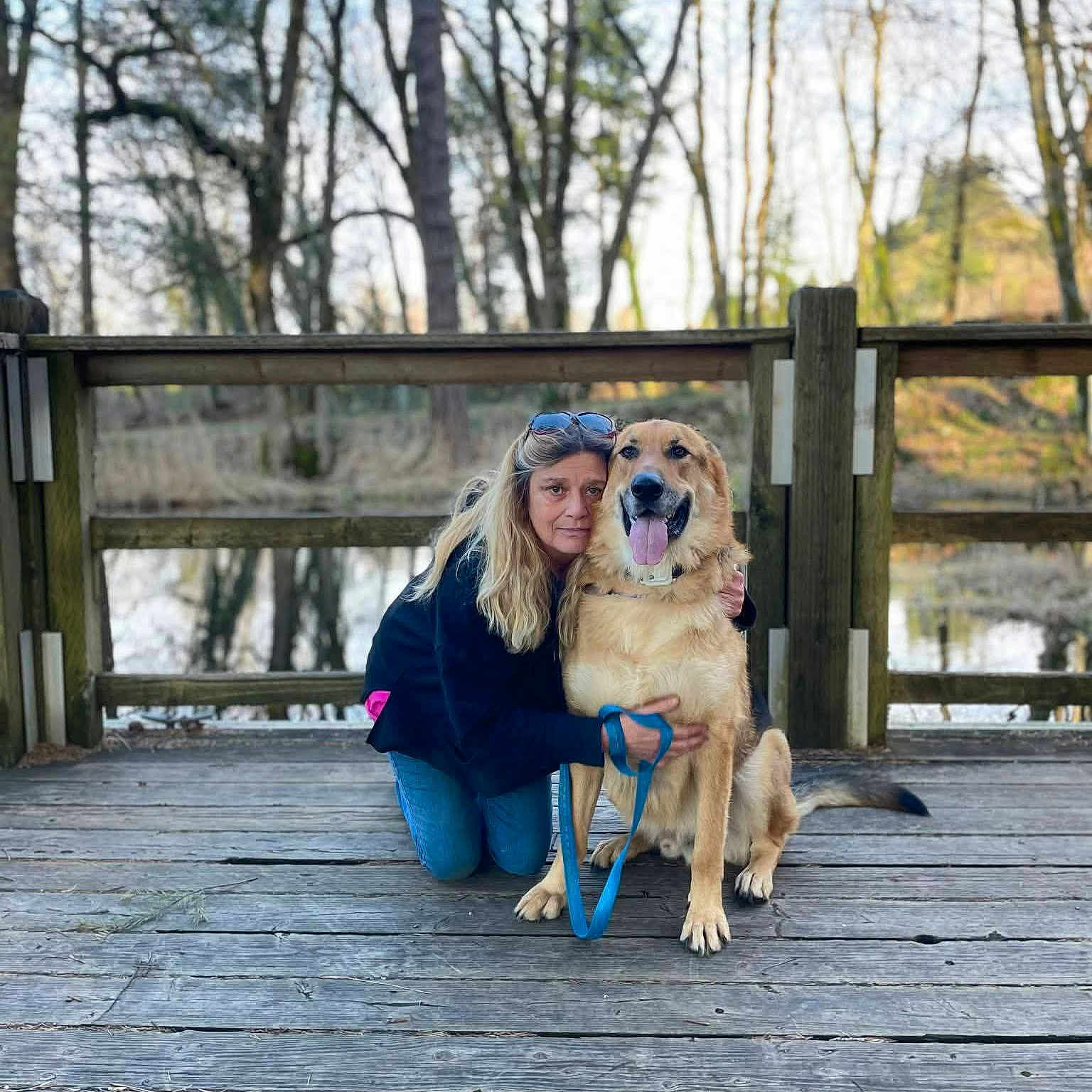 Boomer is registered to the contest to win money with this photo: woman, dog, wooden_dock, forest, trees, pond, leash, outdoor, nature, pet, canine, happy, tongue_out, casual_clothing, long_hair, sunlight, reflection, fence, kneeling, companion