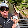adult, baby, casual_clothing, child, greenery, hat, holding, nature, outdoor, park, playful, railings, selfie, smiling, stone_pathway, sunglasses, sunny_day, tongue_out, trees, water_canal
