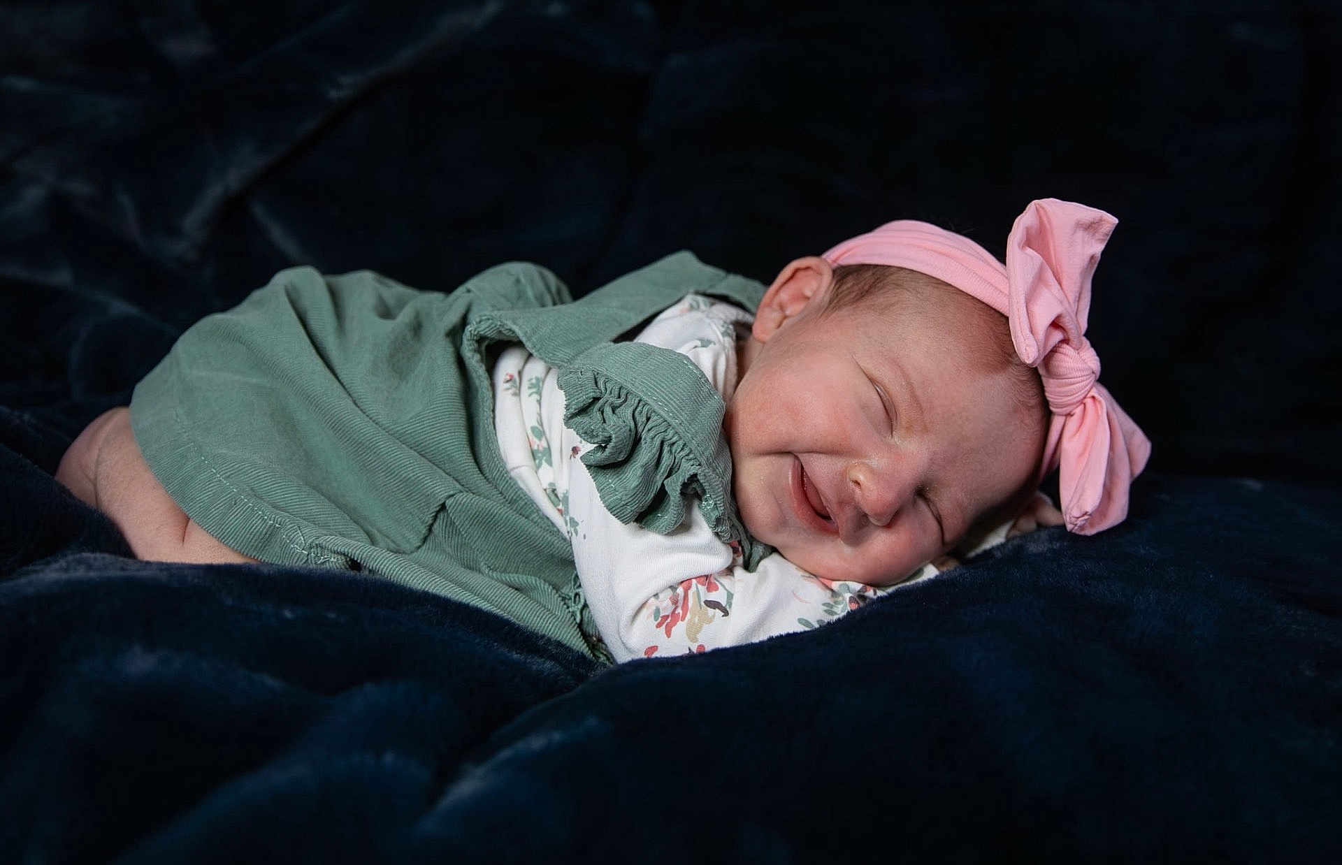 Olive is registered to the contest to win money with this photo: baby, newborn, smiling, sleeping, pink_bow, headband, green_dress, floral_pattern, soft_blanket, dark_background, peaceful, cute, infant, child, portrait, indoors, closeup, resting, happy, toddler
