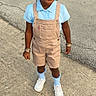 child, smiling, portrait, outdoor, standing, pavement, street, overalls, shirt, braids, hair_ribbons, socks, sneakers, bracelets, fashion, daylight, full_body, person, happy, casual_clothing
