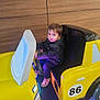 child, cute, floor, fun, indoor, jacket, kid, lighting, number_86, play, portrait, seat, seat_belt, sneakers, steering_wheel, toddler, toy_car, vehicle, wooden_wall, yellow_car