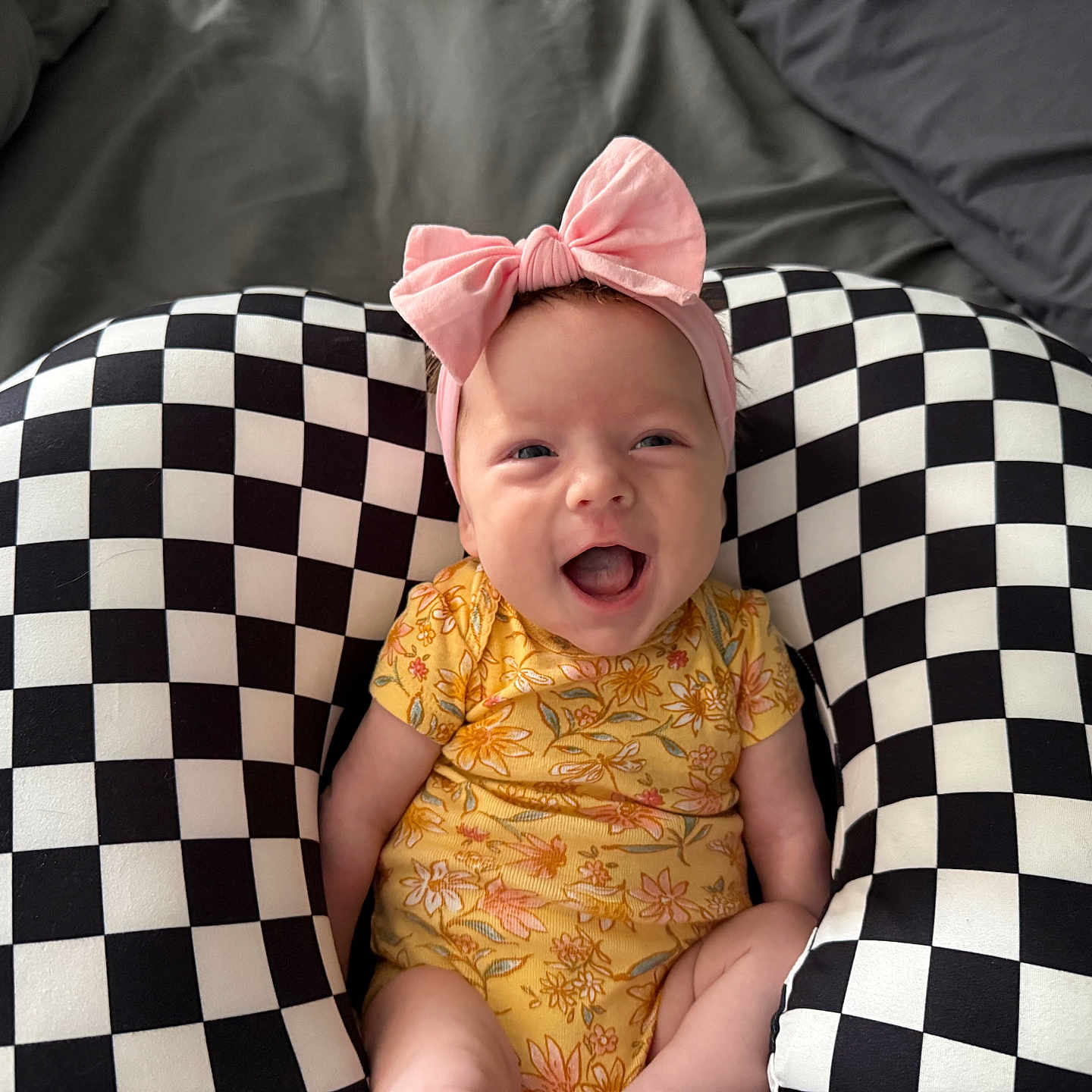 Skylan joined the competition — help win amazing prizes! baby, bed, black_and_white, blanket, checkered_pillow, cute, expression, fabric, happy, headband, indoor, infant, newborn, pattern, pink_bow, portrait, sitting, smile, support_pillow, yellow_onesie