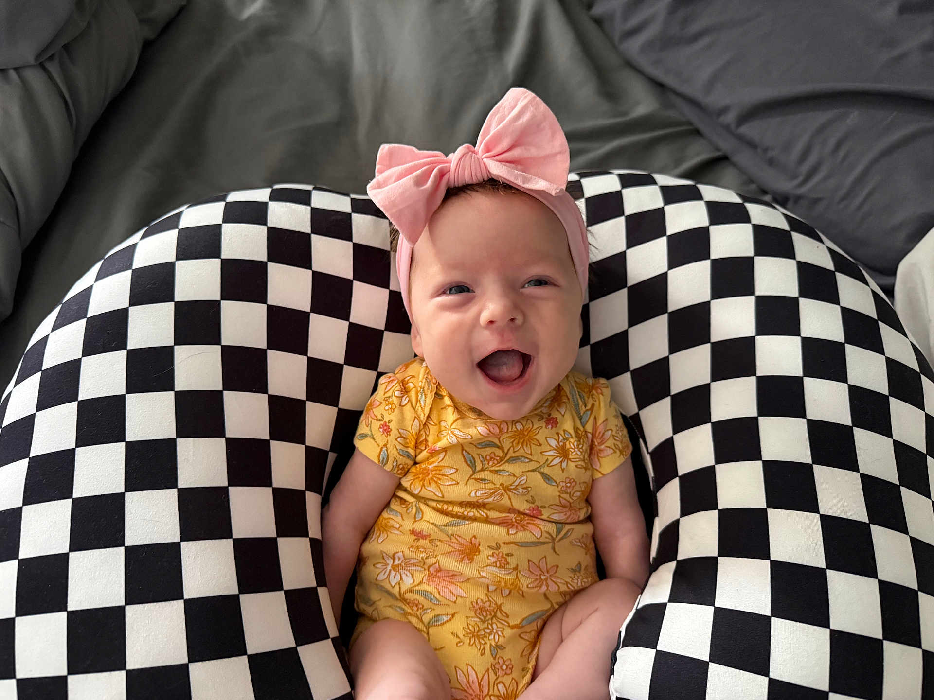 Skylan joined the competition — help win amazing prizes! baby, infant, smile, pink_bow, headband, yellow_onesie, checkered_pillow, black_and_white, pattern, bed, blanket, happy, cute, portrait, indoor, newborn, sitting, expression, support_pillow, fabric