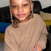 Inaaya participe au concours pour gagner de l'argent avec cette photo : child, braided_hair, top_bun, beige_sweater, fuzzy_sweater, smiling, indoor, couch, cushions, black_leather, person, holding_object, blue_object, orange_object, young_child, portrait, casual_clothing, seated, cozy, home