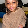 child, braided_hair, top_bun, beige_sweater, fuzzy_sweater, smiling, indoor, couch, cushions, black_leather, person, holding_object, blue_object, orange_object, young_child, portrait, casual_clothing, seated, cozy, home