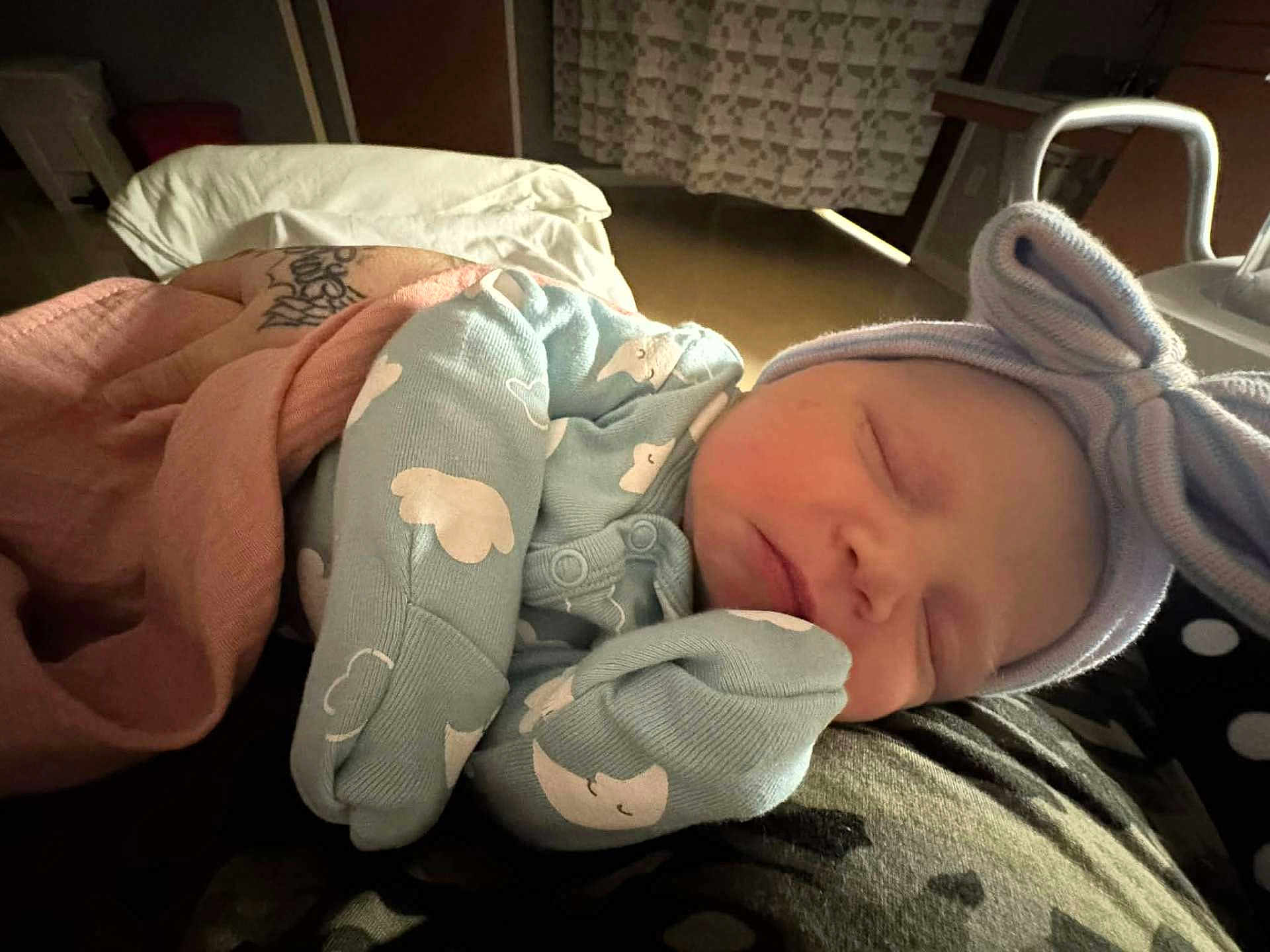 Azariah is registered to the contest to win money with this photo: baby, newborn, sleeping, onesie, hat, bow, cloud_pattern, blanket, tattoo, arm, indoor, resting, cozy, soft_lighting, person, infant, cute, peaceful, clothing, nap