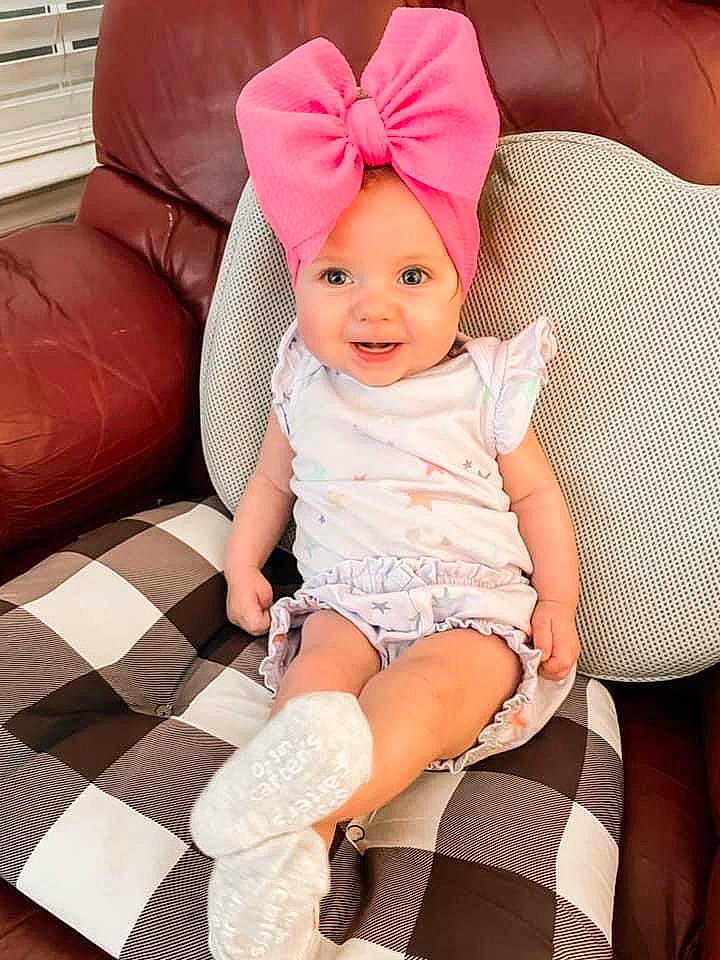 Charley is registered to the contest to win money with this photo: baby, baby_toddler_clothing, cap, comfort, costume_hat, couch, eye, happy, headgear, headwear, knee, lap, leg, pattern, person, pink, purple, sleeve, smile, textile
