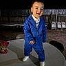 toddler, child, blue_suit, white_shoes, table, outdoor, night, smiling, happy, fence, chairs, stacked_chairs, formal_wear, person, standing, joyful, expression, playful, ground, party
