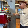 toddler, child, hat, straw_hat, candy, candy_dispenser, pointing, curious, indoor, clothing, calvin_klein, face, person, toy, colorful, blurred_background, floor, jeans, short_sleeve, focus