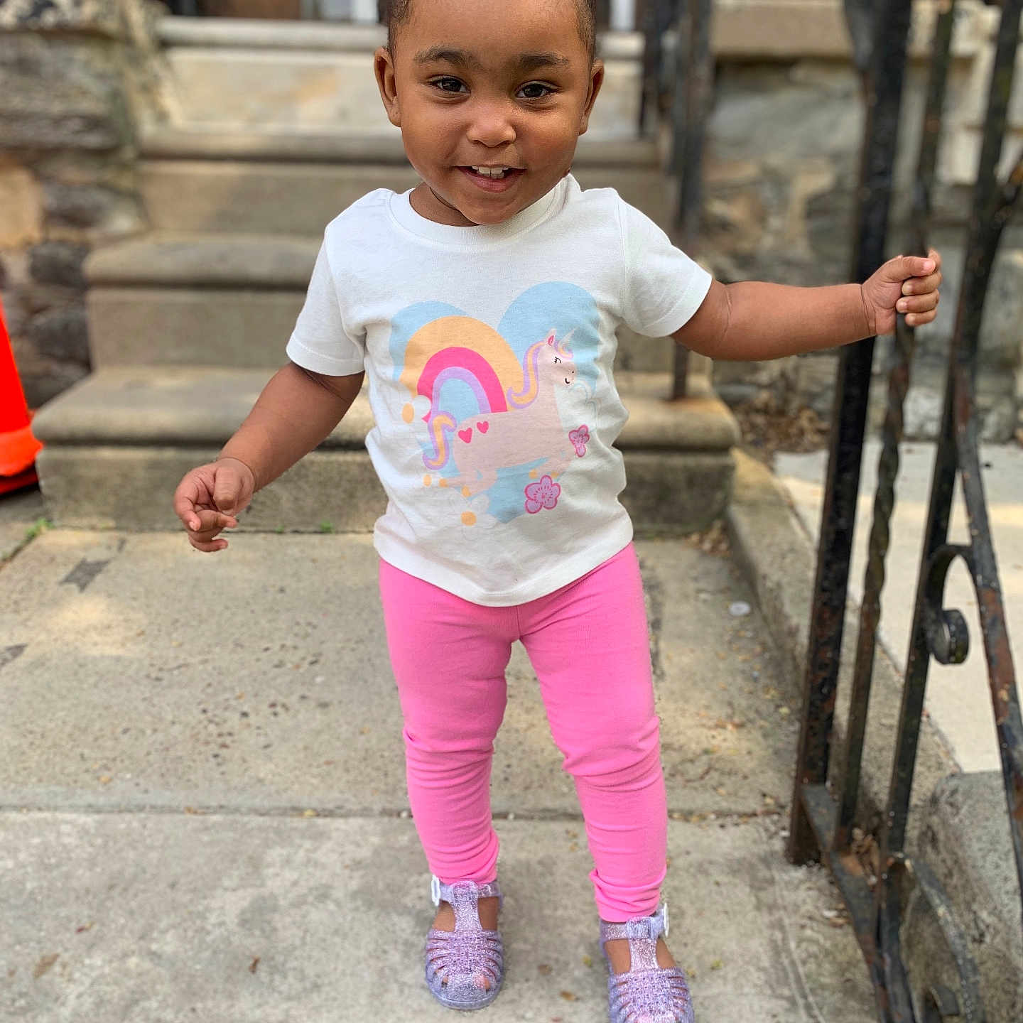 Ahzuri is registered to the contest to win money with this photo: casual_clothing, child, concrete, curly_hair, daylight, girl, handrail, happy, outdoor, pink_leggings, playful, purple_shoes, rainbow, sidewalk, smiling, stairs, standing, toddler, unicorn_print, white_shirt