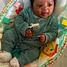 baby, infant, child, green_outfit, pacifier, cushion, colorful, pattern, smiling, tongue_out, cozy, soft_texture, playful, cute, indoor, seated, toy, happy, person, portrait
