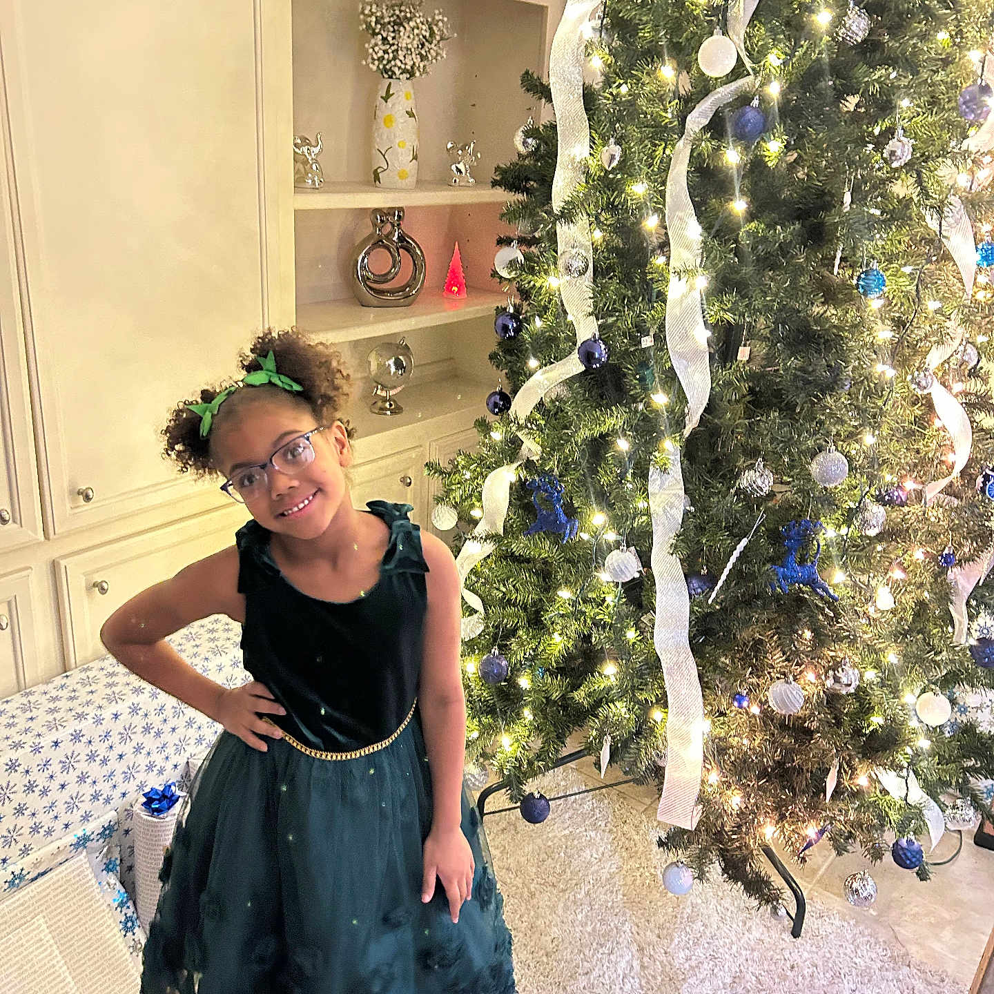 Khristyna is registered to the contest to win money with this photo: cabinet, carpet, celebration, child, christmas_tree, decor, festive, girl, glasses, green_dress, holiday, home_interior, lights, ornaments, pose, presents, ribbon, smile, vase, wrapped_gifts