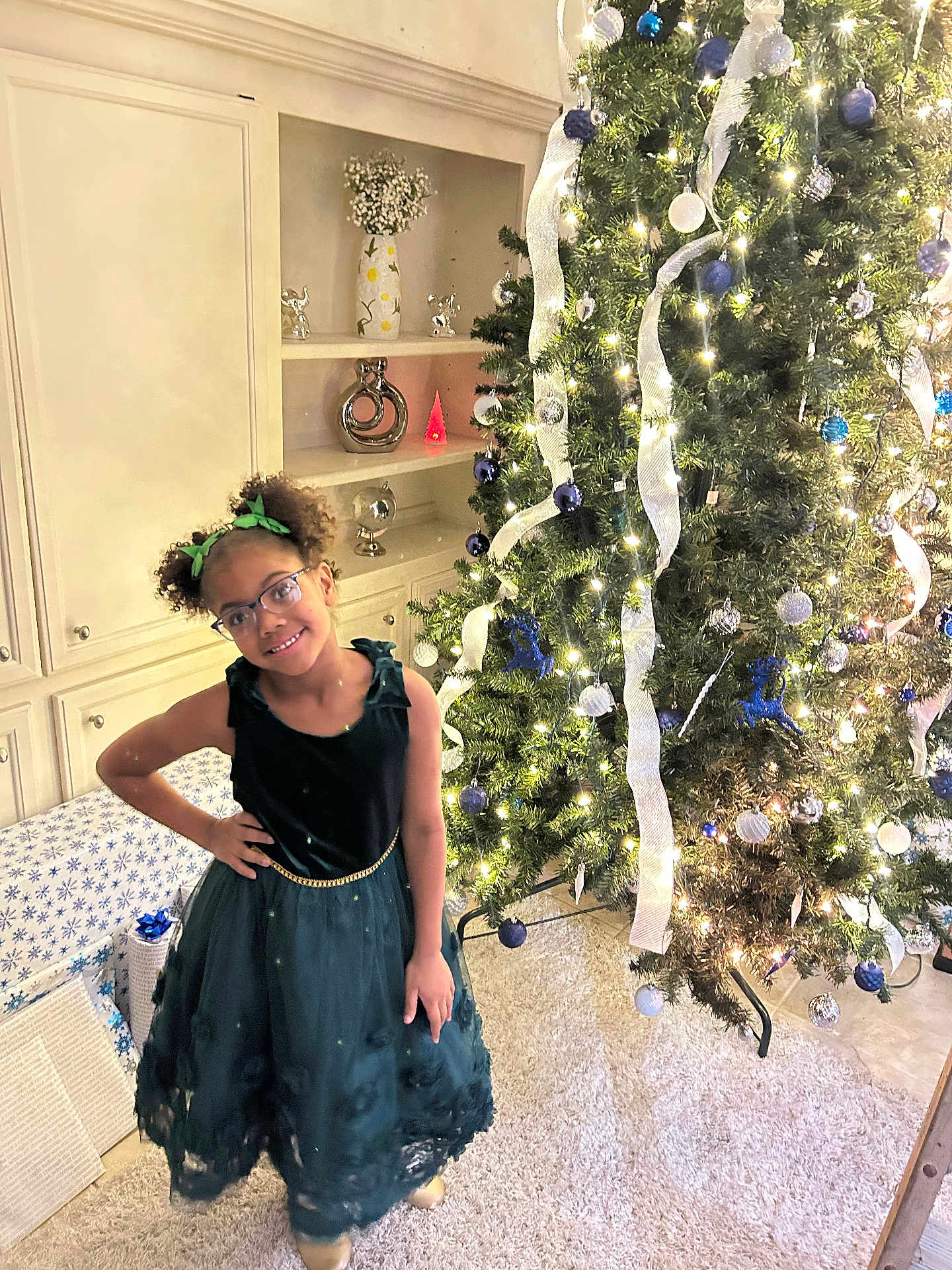 Khristyna is registered to the contest to win money with this photo: cabinet, carpet, celebration, child, christmas_tree, decor, festive, girl, glasses, green_dress, holiday, home_interior, lights, ornaments, pose, presents, ribbon, smile, vase, wrapped_gifts