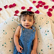 Shiviyan is registered to the contest to win money with this photo: baby, denim_overalls, sunglasses, rose_petals, cushion, star_pattern, blanket, infant, cute, smiling, indoors, portrait, child, relaxing, fashion, adorable, person, headwear, soft_texture, casual