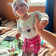 Alice joined the competition — help win amazing prizes! baby, infant, child, portrait, standing, indoors, bedroom, bed, blanket, window, sunlight, hat, cat_ears, pajamas, cupcakes, plaid_pants, socks, adult_hand, tablet, bottle