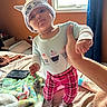 baby, infant, child, portrait, standing, indoors, bedroom, bed, blanket, window, sunlight, hat, cat_ears, pajamas, cupcakes, plaid_pants, socks, adult_hand, tablet, bottle