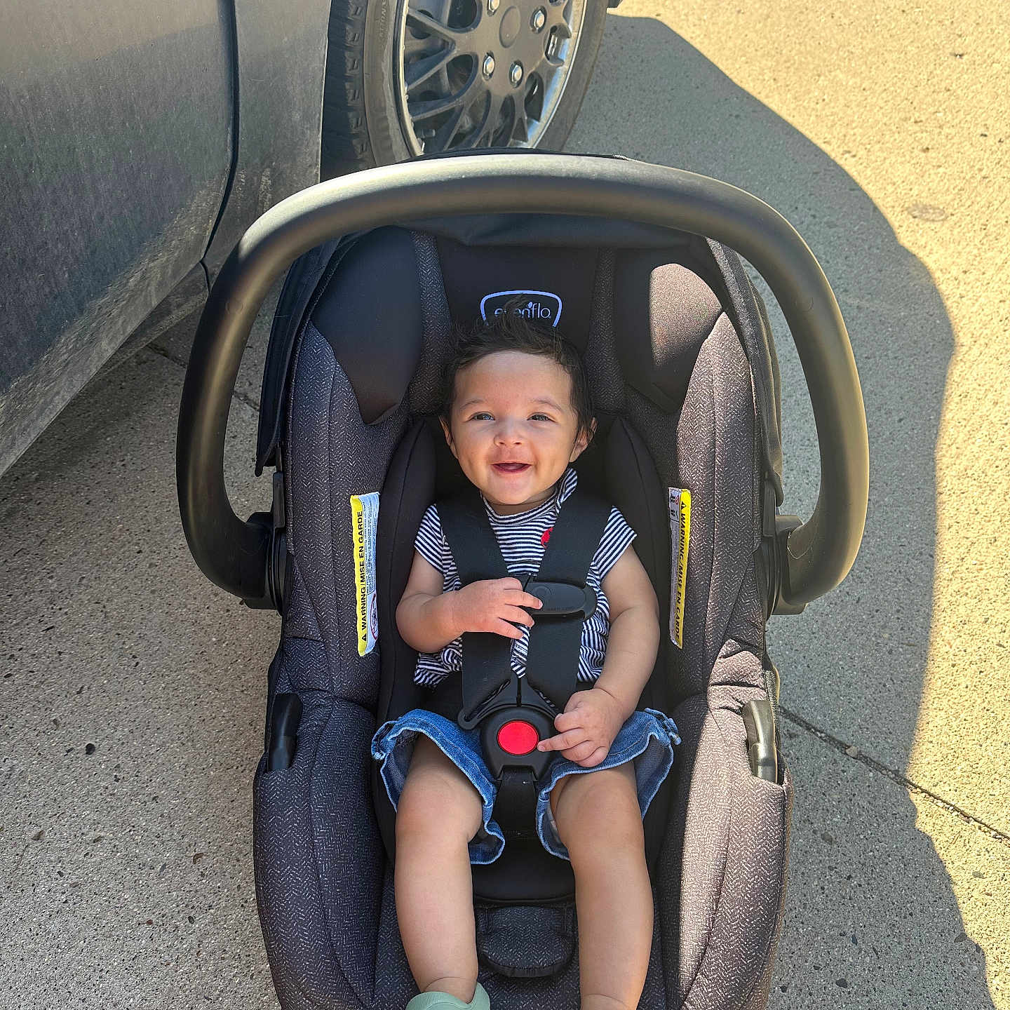 Alice joined the competition — help win amazing prizes! baby, child, infant, car_seat, stroller, smiling, outdoor, parked_car, vehicle, safety_harness, striped_shirt, denim_shorts, green_shoes, sunlight, shadow, pavement, portrait, sitting, happy, automotive
