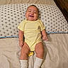 baby, bed, bedding, bedroom, child, face, happy, indoors, infant, laughing, lying_down, onesie, pacifier, patterned_blanket, photography, pillow, portrait, smiling, socks, yellow_outfit