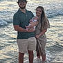 family, baby, man, woman, ocean, water, waves, sunset, beach, pacifier, holding, smiling, casual_clothing, shorts, dress, glasses, hat, outdoor, nature, foot_in_water