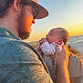 man, baby, beach, sunset, ocean, hat, glasses, facial_hair, holding, sky, nature, outdoor, person, clothing, love, affection, warm_light, portrait, family, happy