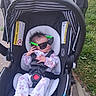 accessory, baby, car_seat, child, clothing, cute, fashion, footwear, grass, green, hair, hand, head, infant, onesie, outdoor, person, safety_harness, sidewalk, sunglasses